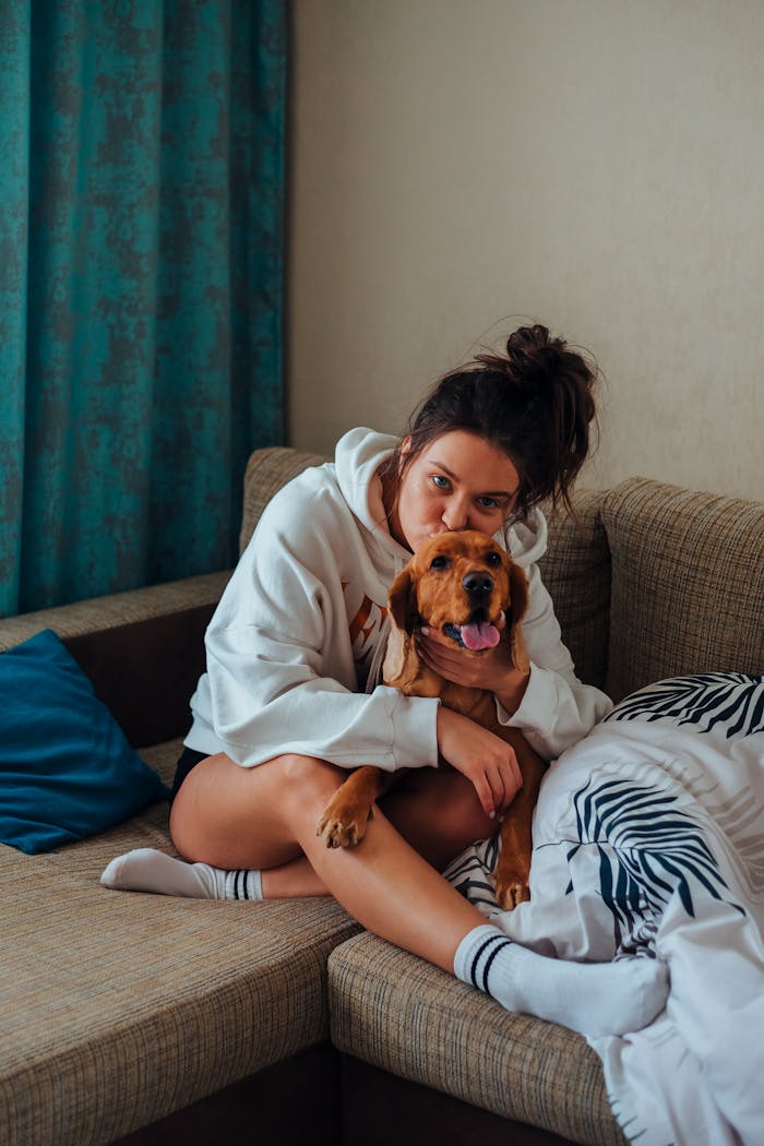 Young woman in hoodie hugging her dog on a comfortable sofa in a warm, cozy living room setting.
